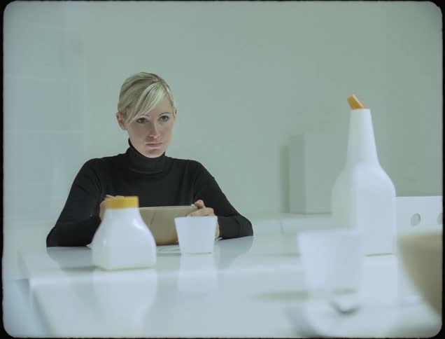 A woman wearing black sits at the end of a white table eating from a large bowl with a large spoon