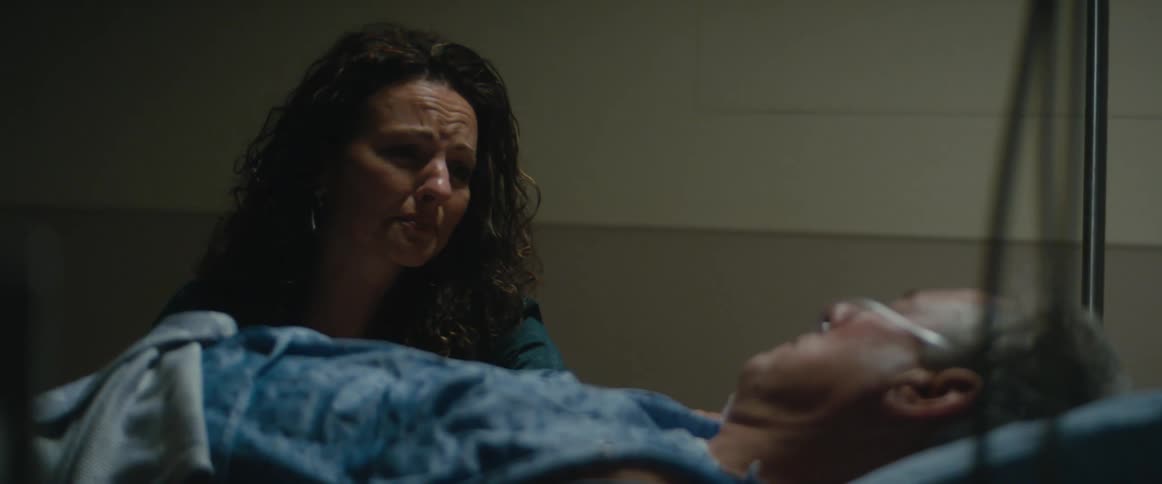 A woman reaches out to touch her dad on a hospital bed her face lined with worry