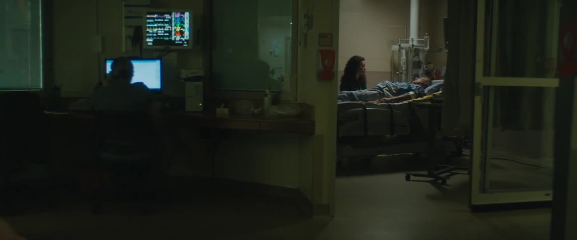 A woman sits by a mans bed in a dimly lit hospital room while a nurse wheels a cart past