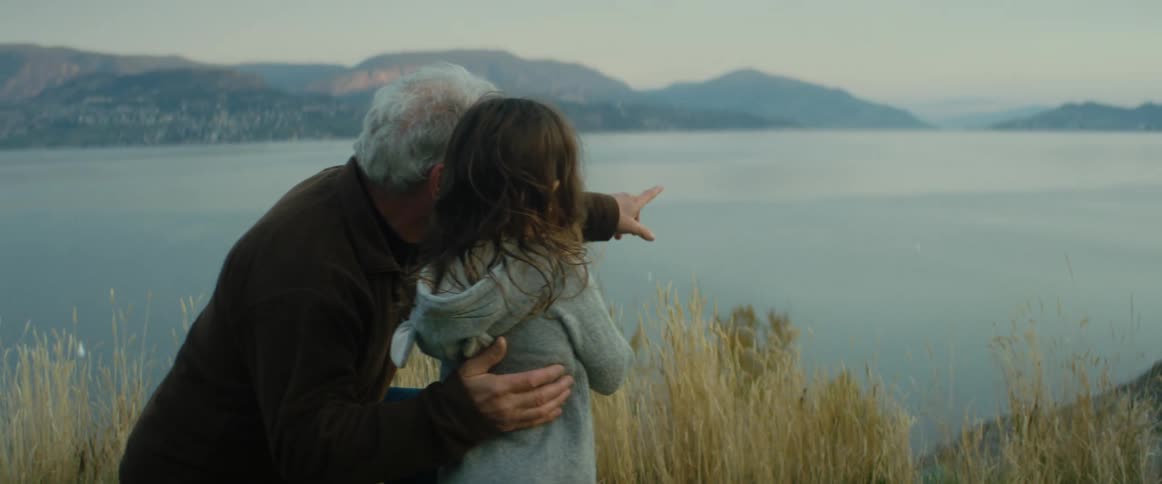 A man kneeling in the grass next to a little girl pointing at the lake below