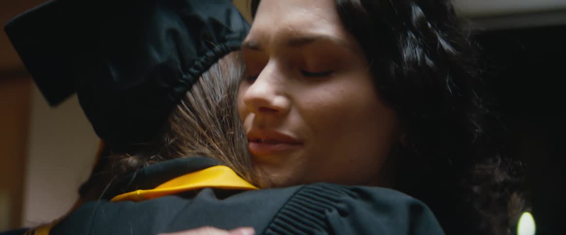 A girl wearing a graduation cap and gown hugs a woman