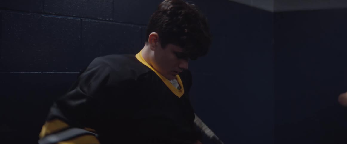 A boy puts on a sports jersey and the other boy is sitting down holding a hockey stick