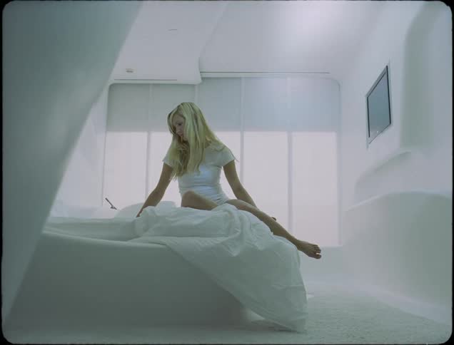 A teenage girl with blond hair is sitting on a bed as the window shade behind her slowly goes up