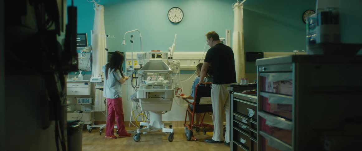 A man wheels a woman into the nicu where she reaches into an incubator to touch her baby