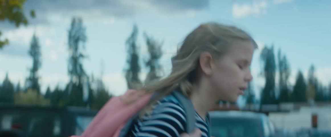 A young girl carrying a backpack runs past several parked cars