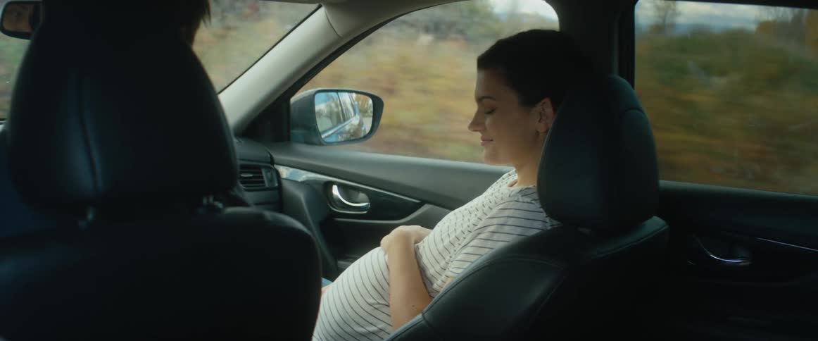 A pregnant woman rides in the passenger seat of a car as it travels down a rural road