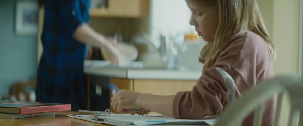 A girl is sitting at a kitchen table doing her homework as someone washes dishes in the sink