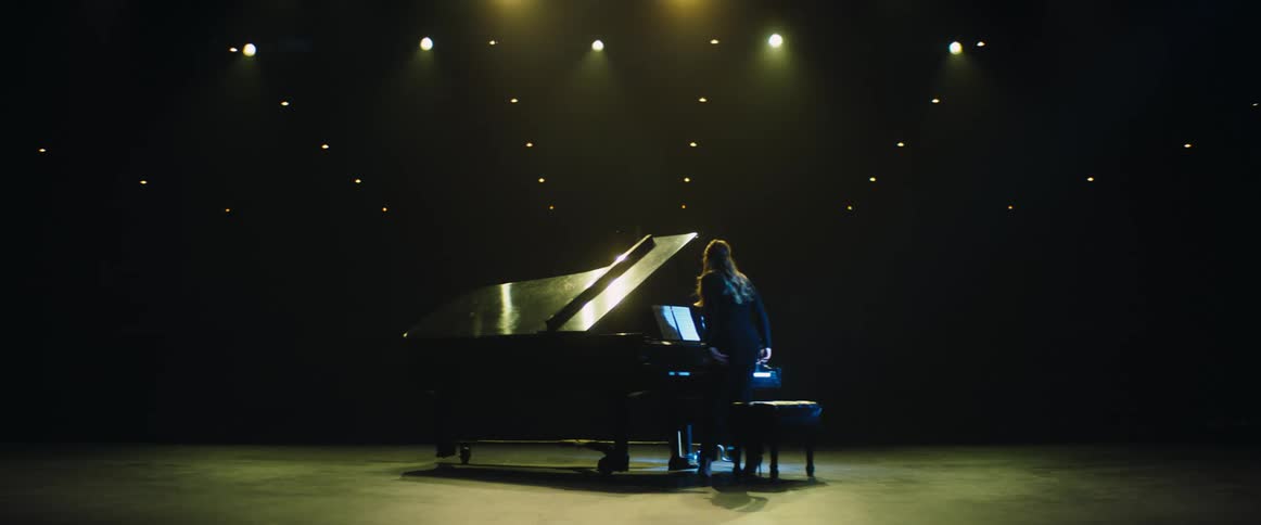 A girl walks on stage and sits down and starts playing a grand piano