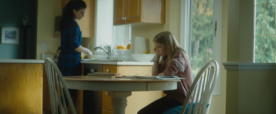 Mom washes dishes at the sink while her daughter looks bored and frustrated doing schoolwork