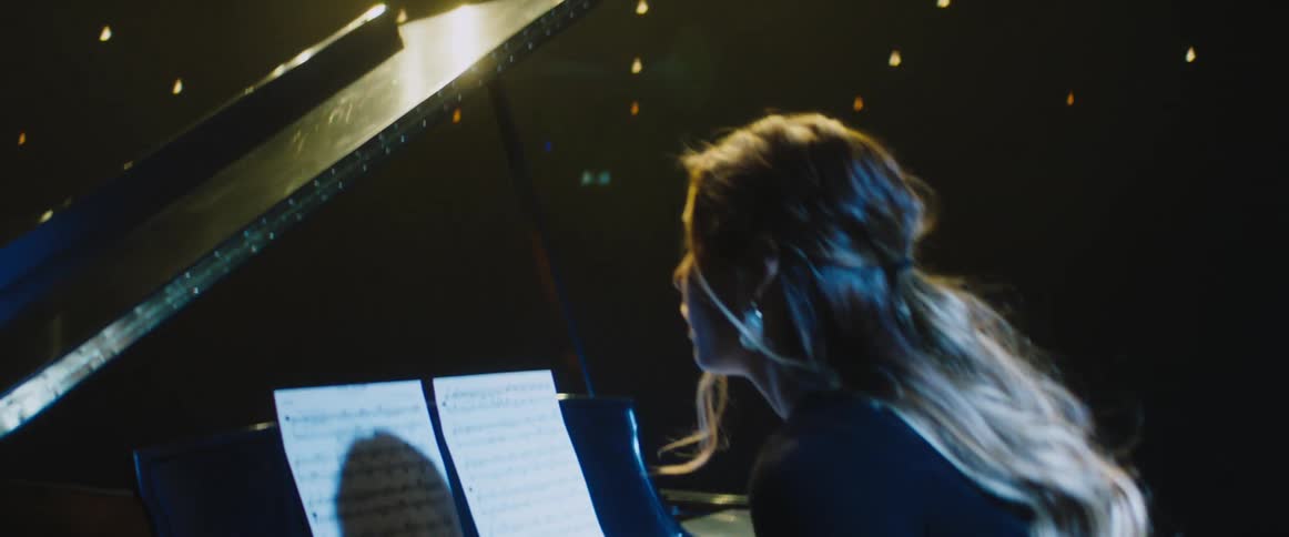 A woman walks on stage and sits down at the piano adjusting her sheet music