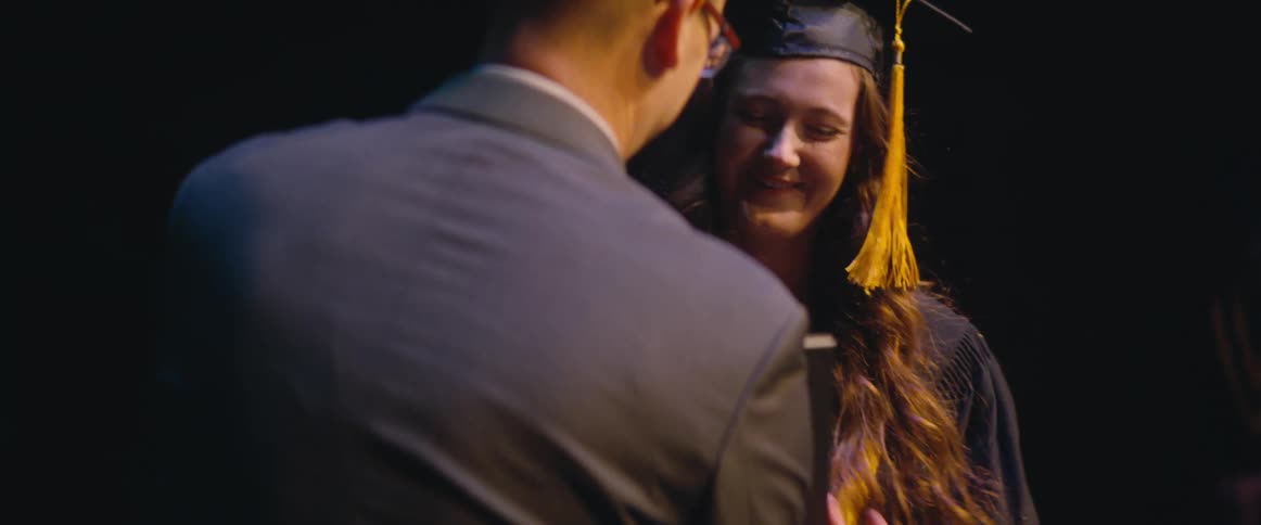 A woman wearing a graduation cap and gown walks across the stage and a man shakes her hand as he hands her a diploma