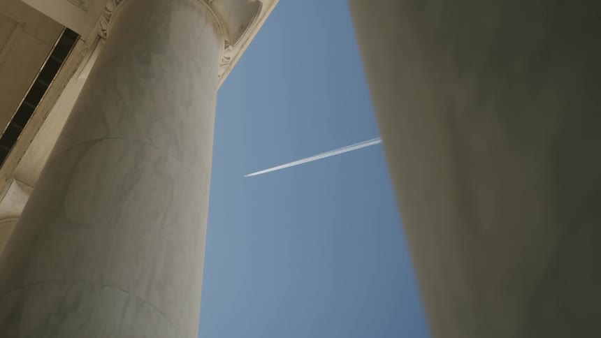 A jet plane flying in a clear blue sky leaving a contrail behind it can be seen between the columns of buildings