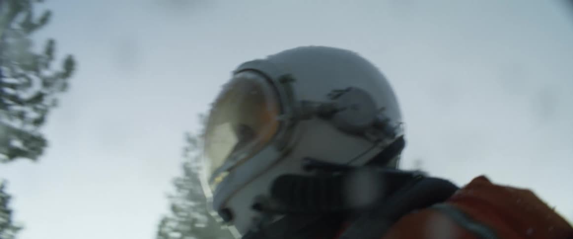 Male pilot wearing flight suit and helmet walks through conifer trees with mountain in background