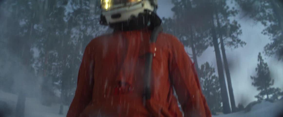 A person stands in a snowy forest wearing a helmet and orange jumpsuit