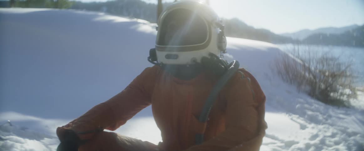 A man wearing a helmet is sitting outside as it snows and there are mountains in the distance