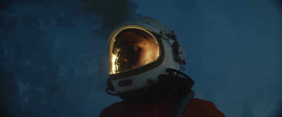 Man wearing pilot crash suit lifts helmet glass to look around before moving across snow covered forest area