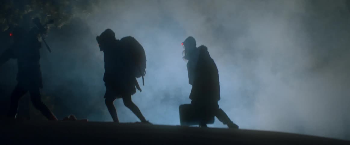 A group of people carry bags and equipment through a smoky forest