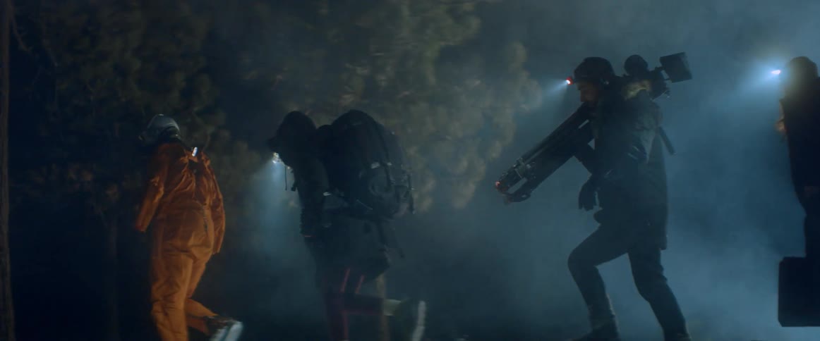 Film crew carrying equipment walks through forest at night along with person wearing pilot crash suit