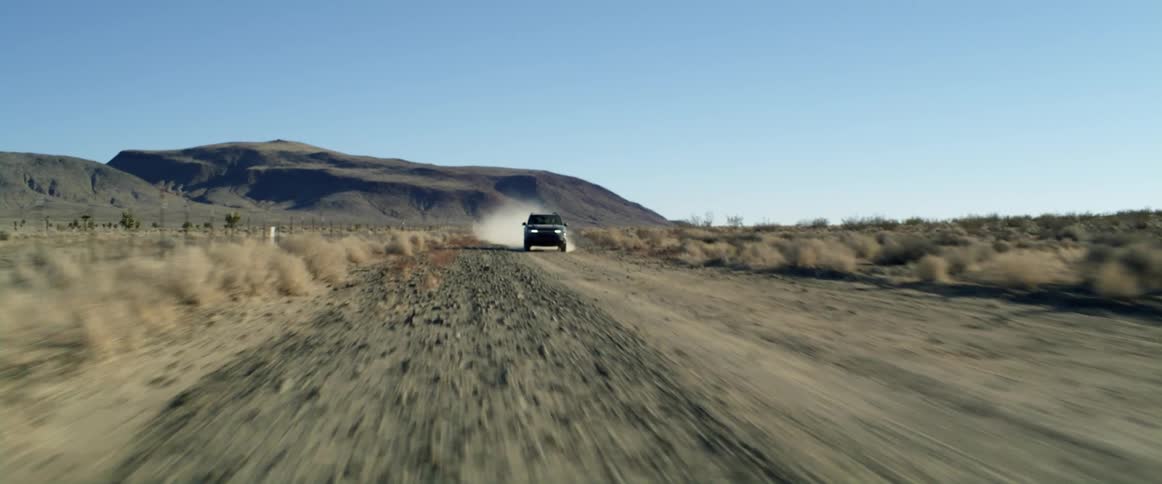Dust flies behind a truck traveling on a desert road with mountains in the distance