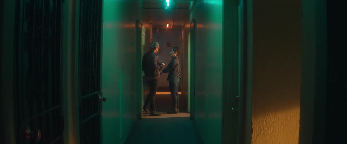 A woman is standing in the hallway of an apartment building as someone opens the door and a man comes out of another door and another man walks down the hall