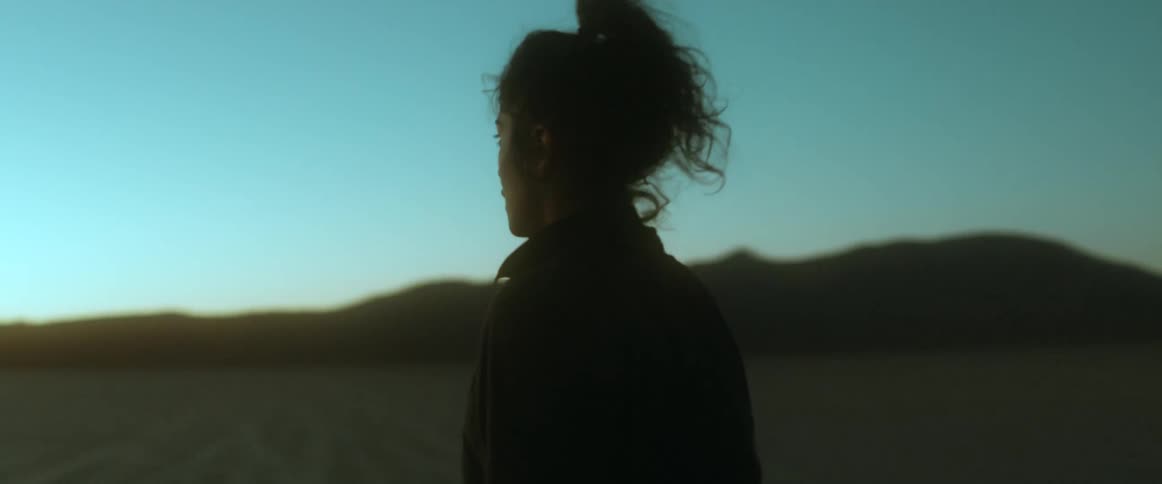 A woman talks to the camera while walking through a desert during the day
