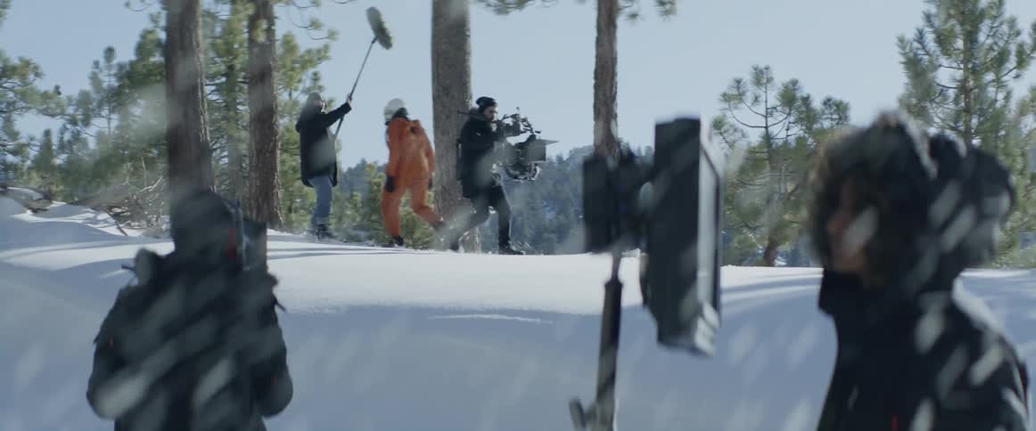 People are outside carrying large items down a hill as it snows and a man is standing in front of a monitor