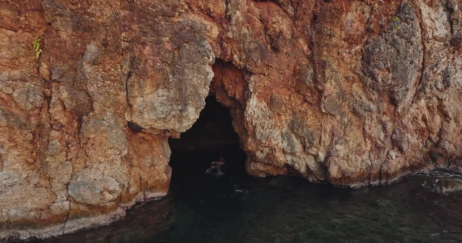 A swimmer swims into a cave at the bottom of a rock face on the ocean during the day