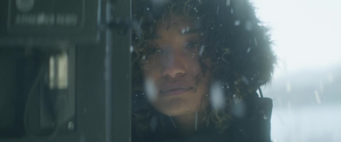 A woman is standing outside near a phone booth wearing a winter coat and she smiles as snow falls