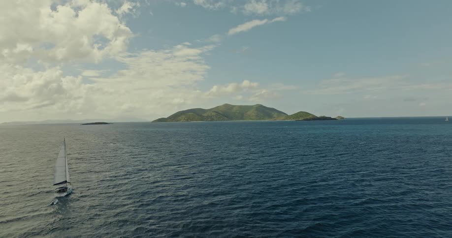 A sail boat travels on the water as it waves and ripples near an island under a cloudy sky