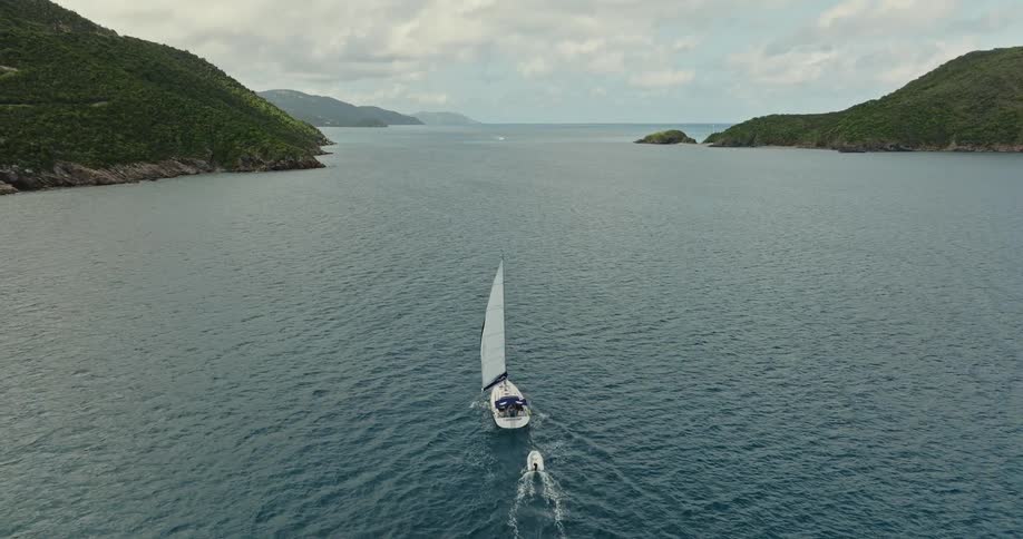 A sail boat travels in the water near shore and there is a smaller boat behind it