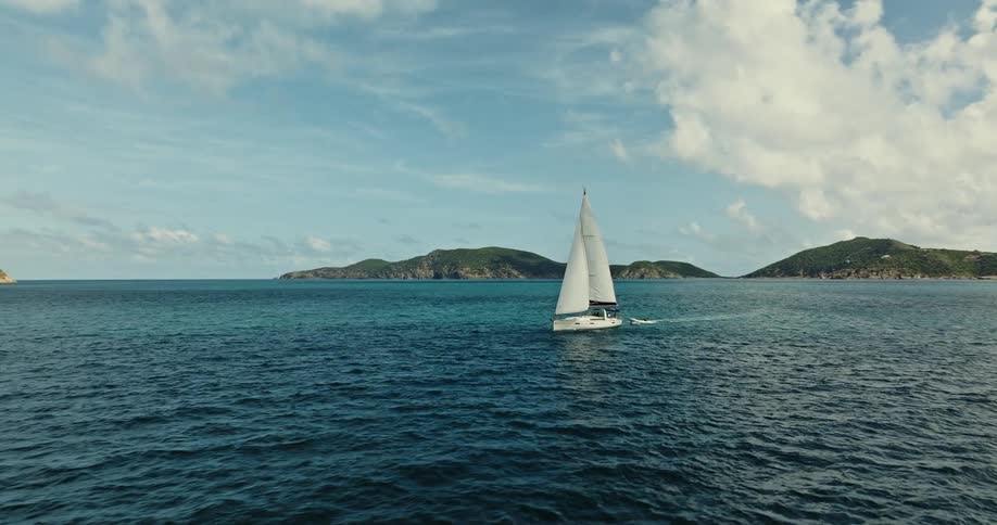A sail boat travels across the ocean while towing a smaller boat behind it