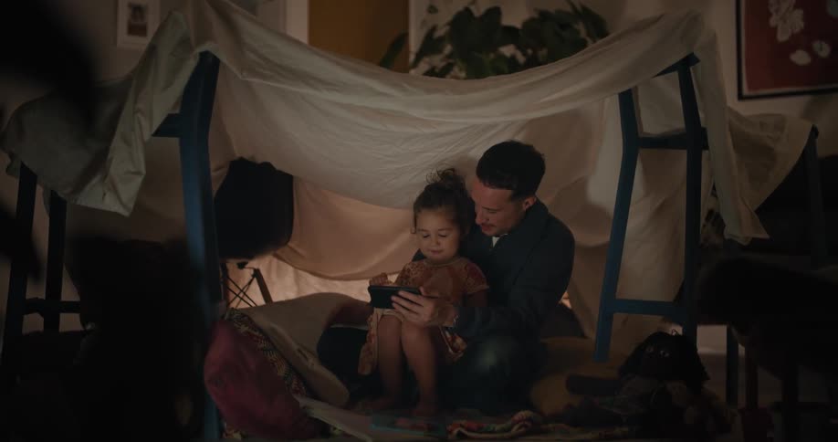 A girl sits in her fathers lap while watching video on a smartphone under a makeshift tent