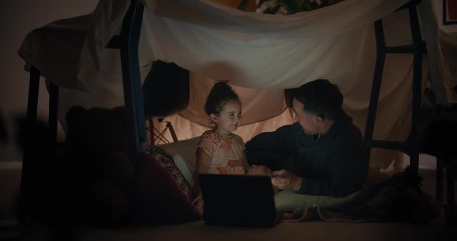 A little girl watches the screen of a laptop with a man in a blanket fort in a dark room