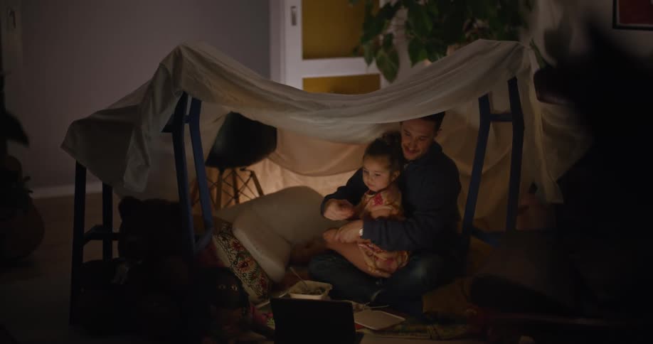 A man hugs his daughter from behind as they sit and watch videos on a laptop while under a fort