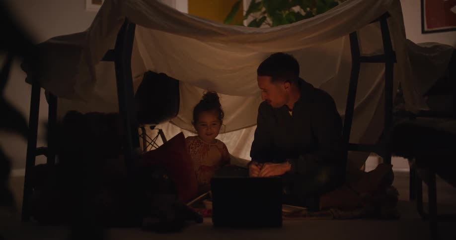A father and daughter camp indoors under a blanket