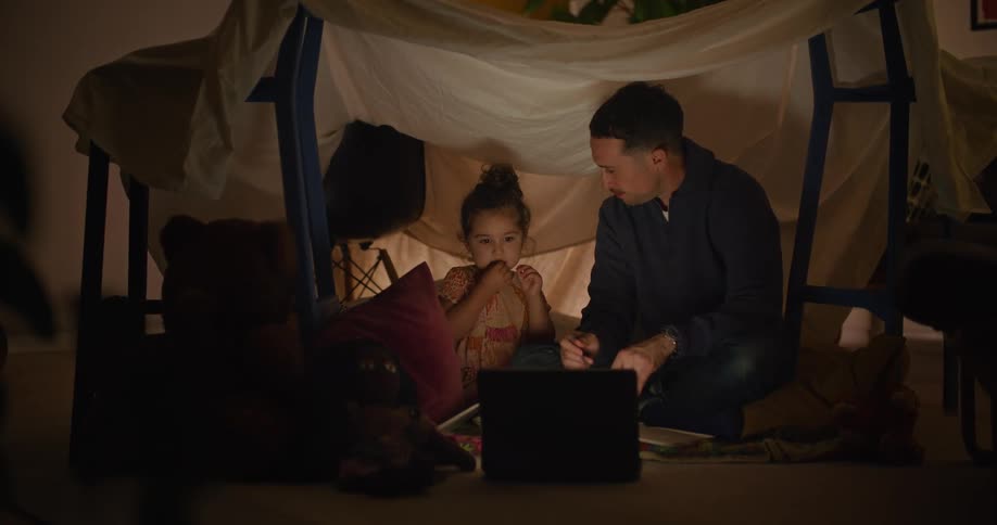 A man and his young daughter are playing in a fort made with chairs and a sheet and they are looking at a laptop computer