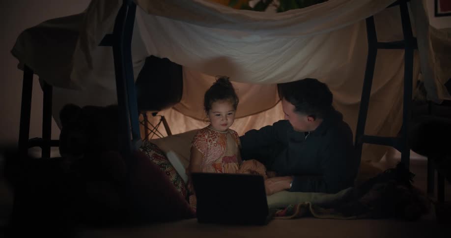 A man and his young daughter are playing in a fort made with chairs and a sheet and looking at a laptop computer