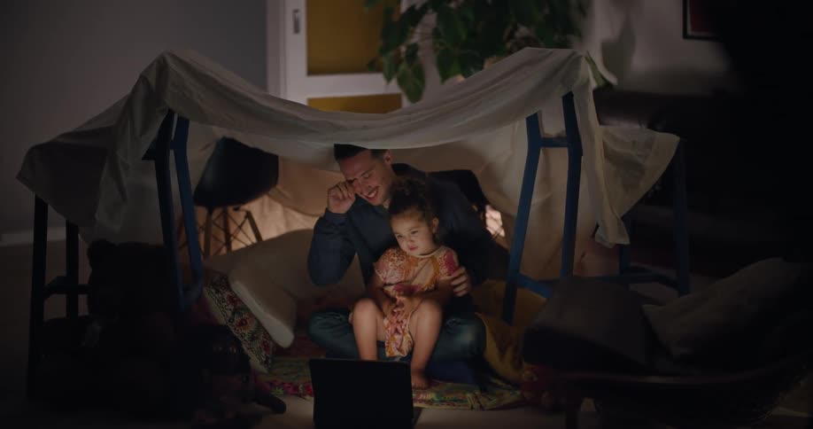 A girl sits in her fathers lap while watching videos on a laptop under a makeshift tent