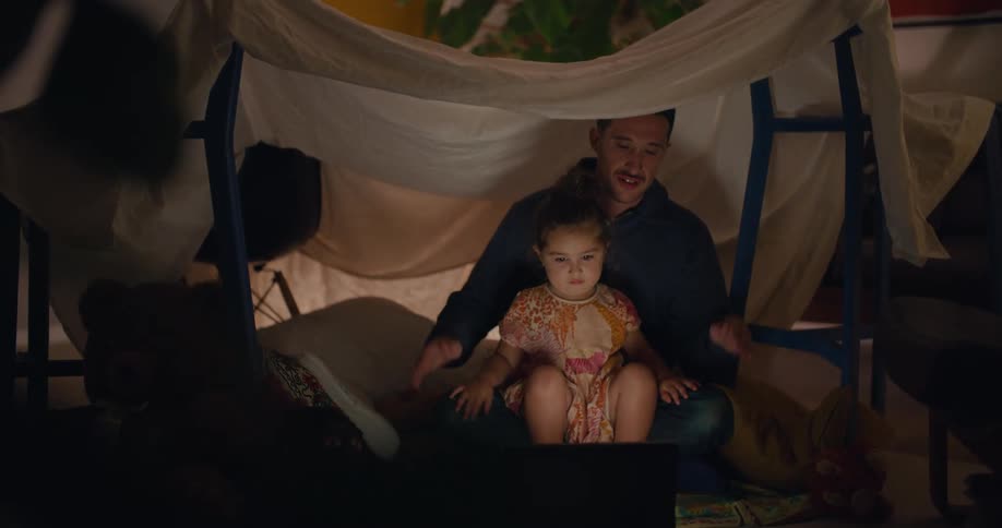 Father and daughter sit under makeshift sheet tent in home and clap while watching a screen