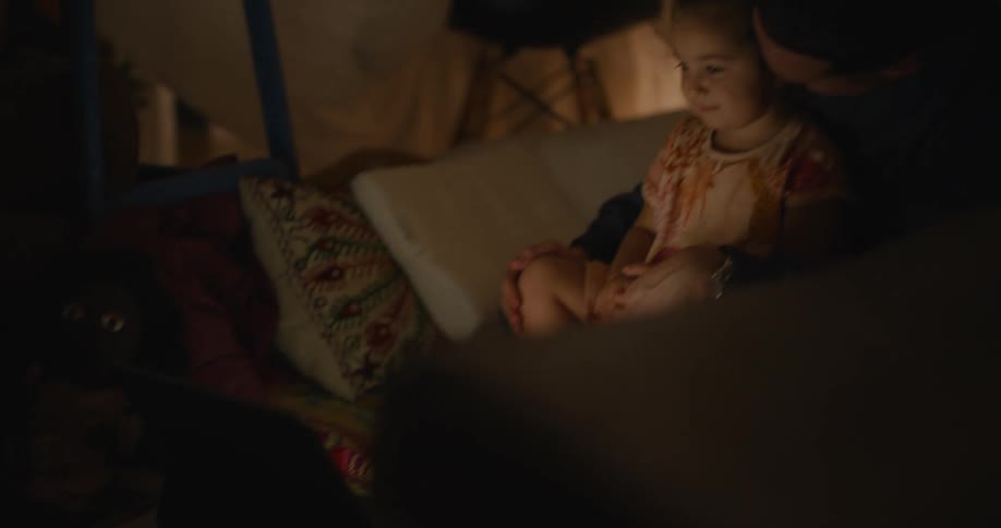 Father and daughter sit near fabric wall viewing video on laptop