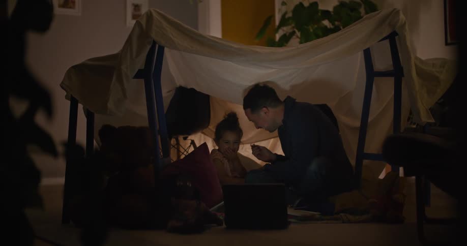 Father feeds daughter as they sit on floor in home under impromptu sheet tent near laptop