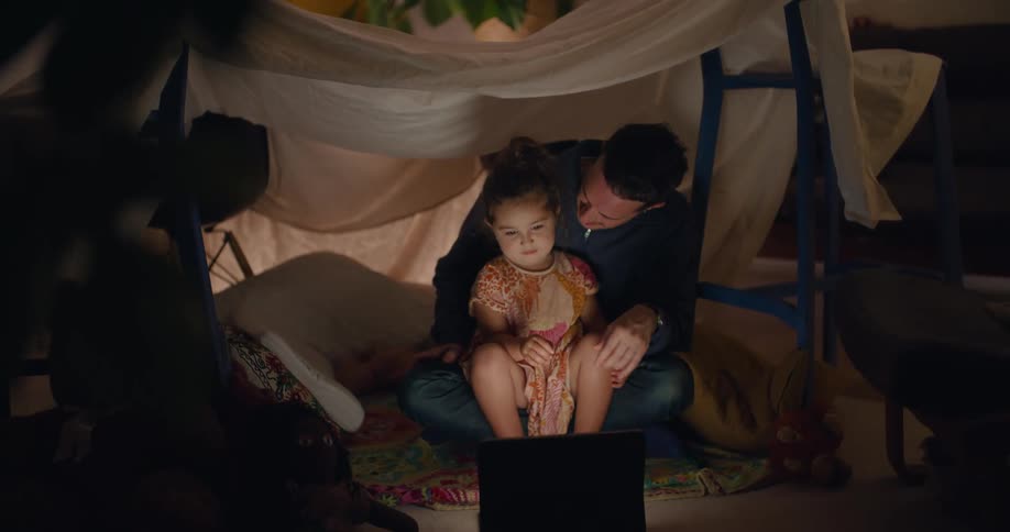 A man and his young daughter are playing in a tent fort made with chairs and a sheet and they are watching something on a laptop computer