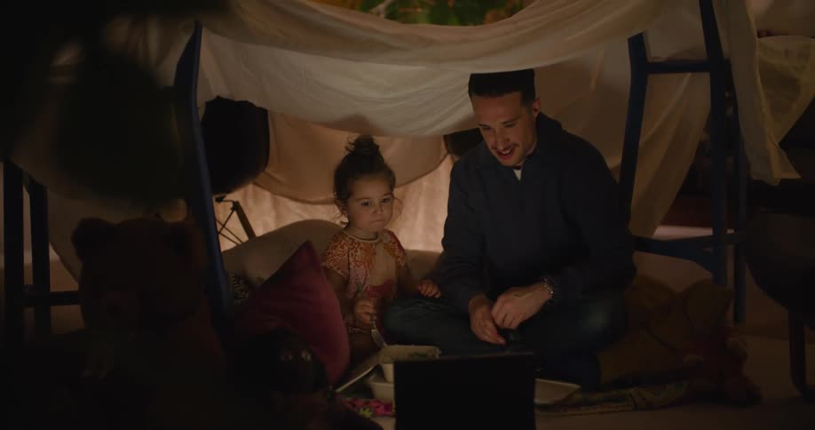 A man and his daughter watch videos on a laptop while camping at home