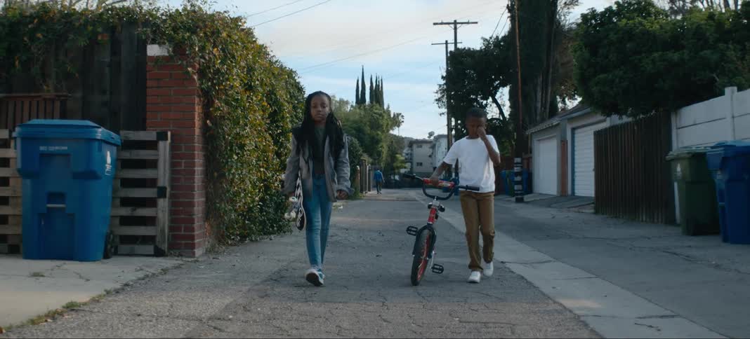 A boy walks his bicycle next to his sister who is carrying a skateboard