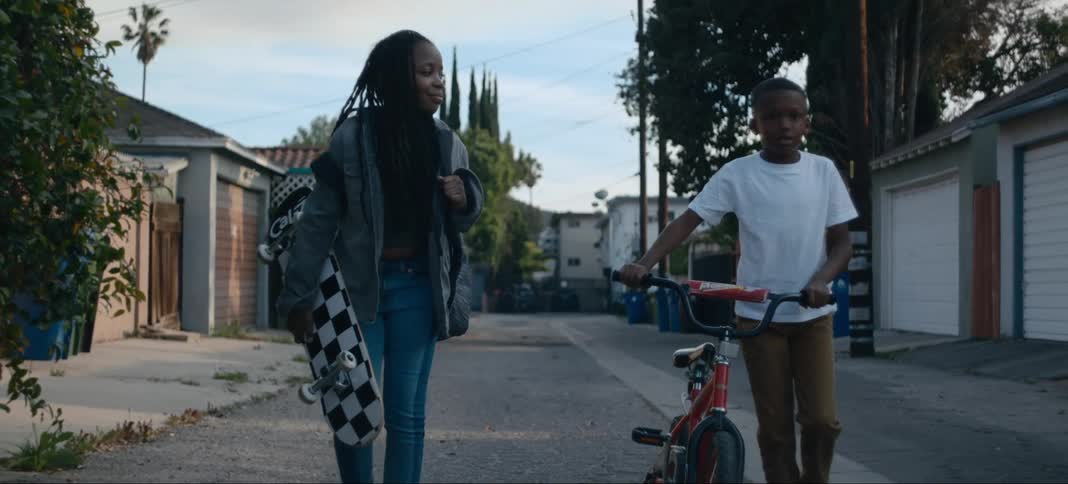 A girl carrying a skateboard walking down a neighborhood street next to a boy wheeling his bike