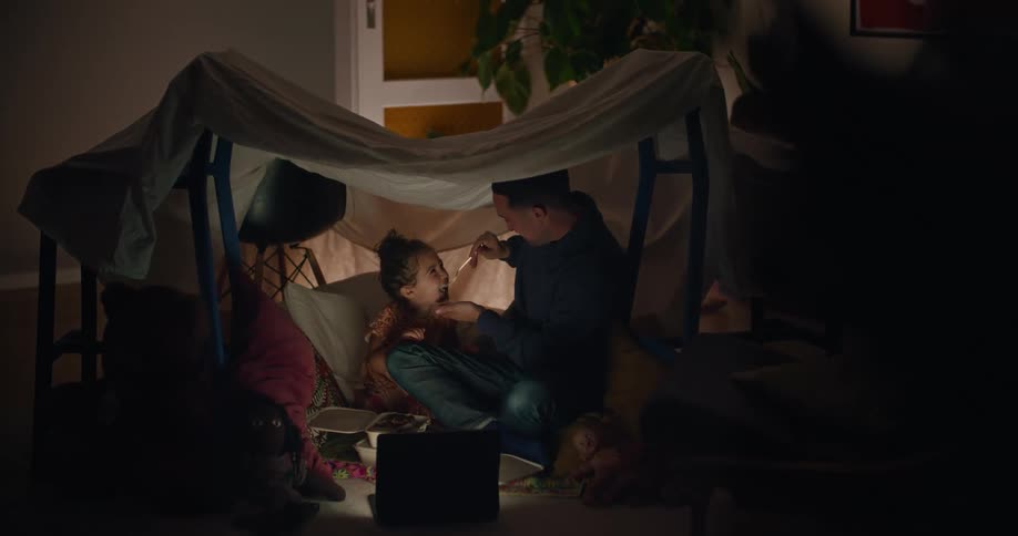 A man and his young daughter are playing in a fort made with chairs and a sheet and he puts a spoonful of food into her mouth