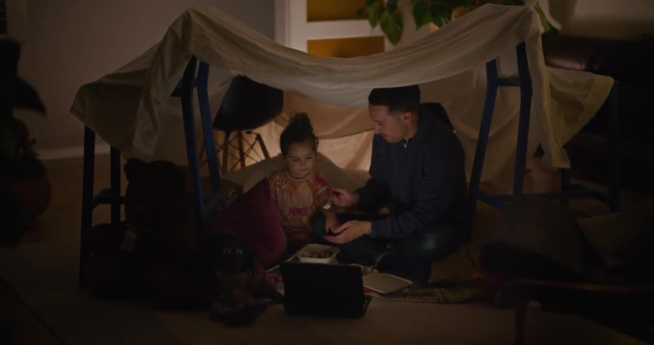 A father feeds his daughter as they sit in a fort at home and watch a laptop