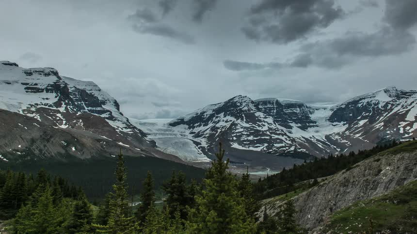 Clouds move over a forest and snowy mountain peaks