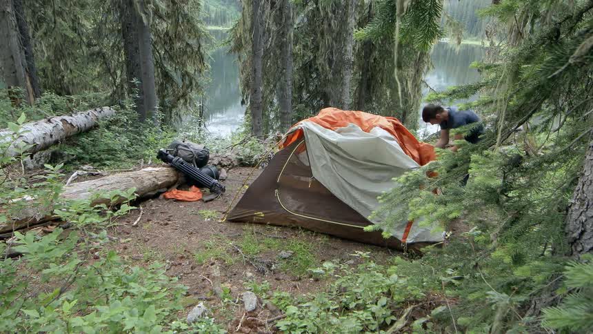 A man sets up a tent in a small clearing by a lake