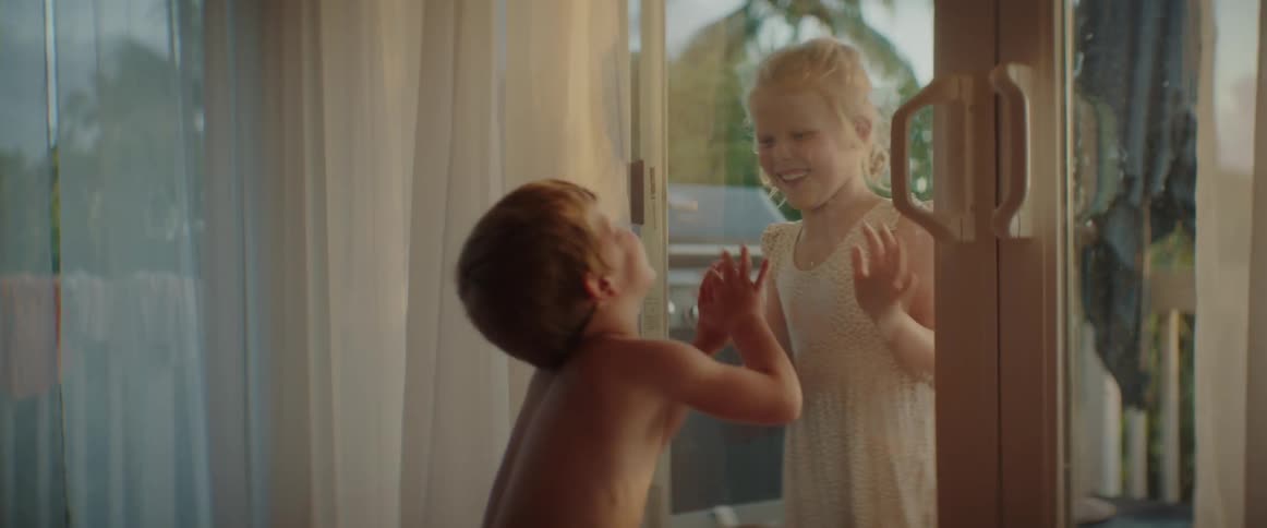 A little boy is inside playing with a girl who is outside and they are putting their hands and faces on a glass door and laughing